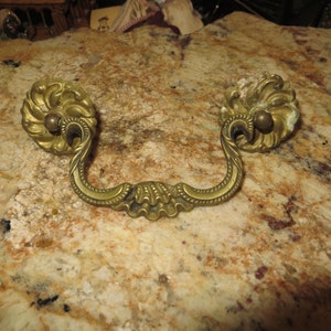 May include: Antique brass drawer pull with ornate floral design and a rounded knob on each end.