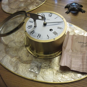 May include: A gold-toned Schatz clock with a white face, black Roman numerals, and black hands. The clock has a magnifying glass attached. A paper with German text is next to the clock. The clock sits on a decorative, gold-colored surface.