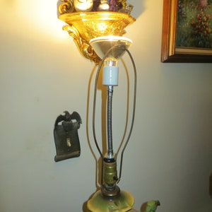 May include: A vintage porcelain lamp with a green and white floral design. The lamp has a flexible metal stem and a white ceramic socket. The lamp is on a white wall with a gold-colored wall sconce above it.