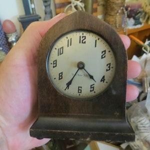 May include: A vintage wooden clock with a white face and black hands. The clock has a simple design with Roman numerals and a small inscription that reads "The Time Teller".