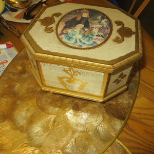 May include: Octagonal wooden box with a gold painted finish and a raised oval image of three girls in vintage clothing. The box is sitting on a gold and white circular tray.