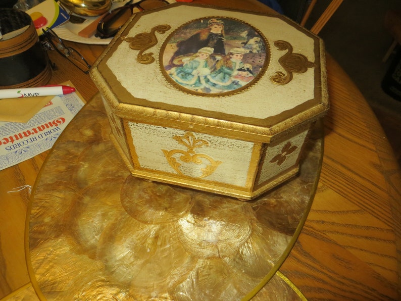 May include: Octagonal wooden box with a gold painted finish and a raised oval image of three girls in vintage clothing. The box is sitting on a gold and white circular tray.