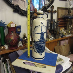 May include: A white ceramic bottle with a blue and white windmill design. The bottle has a spout and a cork stopper. The bottle is sitting on a blue and white surface.