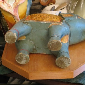 Puede incluir: Una figurita de elefante de madera azul con detalles pintados en oro y amarillo. El elefante est&aacute; acostado de lado con sus patas visibles. La figurita est&aacute; sobre una superficie de madera.