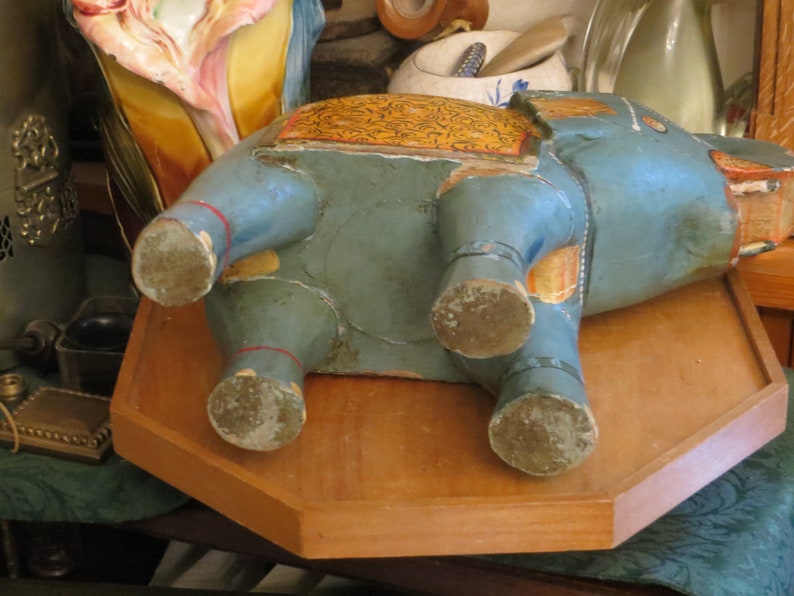 Puede incluir: Una figurita de elefante de madera azul con detalles pintados en oro y amarillo. El elefante est&aacute; acostado de lado con sus patas visibles. La figurita est&aacute; sobre una superficie de madera.