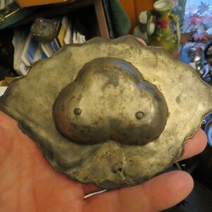 May include: A silver-toned metal trinket dish with a heart-shaped design. The dish has a scalloped edge and two small holes on the heart.