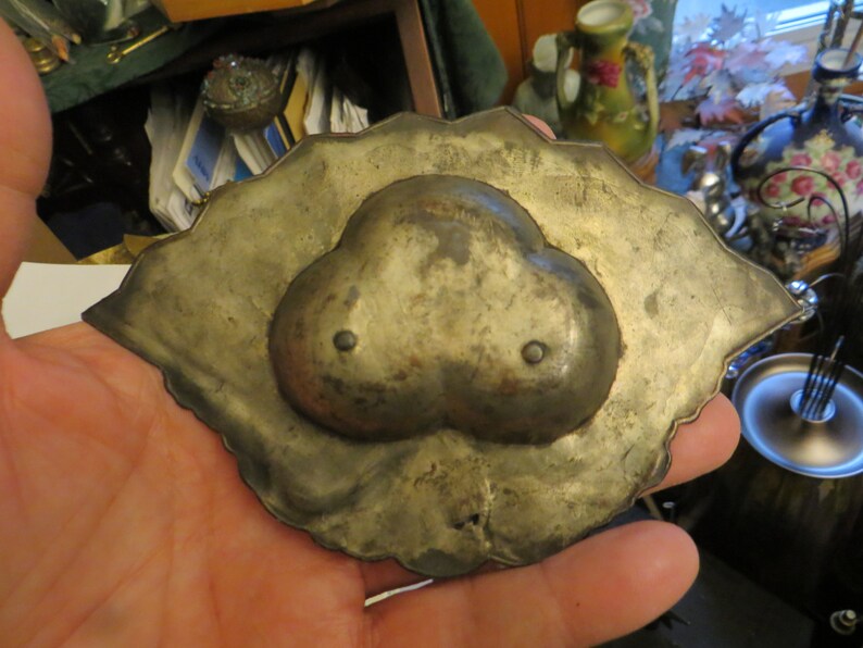 May include: A silver-toned metal trinket dish with a heart-shaped design. The dish has a scalloped edge and two small holes on the heart.