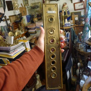 May include: A vintage brass elevator control panel with the words "LIGHT", "ALARM", and "STOP" above several circular buttons. The panel has a rectangular shape and is held by a person. The panel is a golden color.