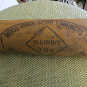 May include: A vintage wooden baseball bat with the text "WOOD RIVER WOOD PRODUCTS CO. ILLINOIS 105" printed in black ink. The bat has a natural wood finish and shows signs of wear.