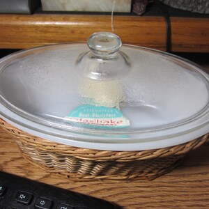 May include: A white glass casserole dish with a wicker basket base and a clear glass lid. The lid has a knob on top and a label that reads "Heat Resistant Glassbake Ovenware".