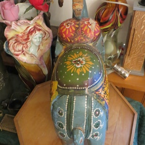 Puede incluir: Una figurita de elefante de madera pintada a mano con pintura azul, verde, amarilla y roja. El elefante est&aacute; de pie sobre una base de madera.
