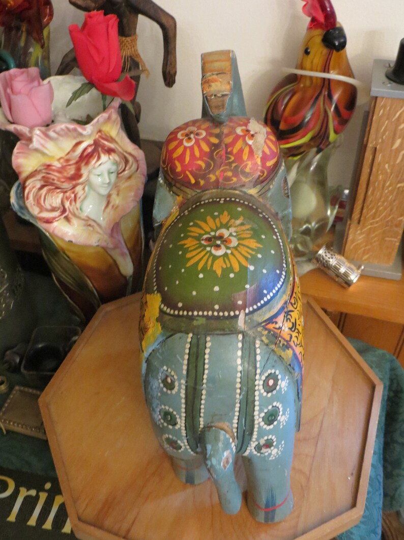 Puede incluir: Una figurita de elefante de madera pintada a mano con pintura azul, verde, amarilla y roja. El elefante est&aacute; de pie sobre una base de madera.