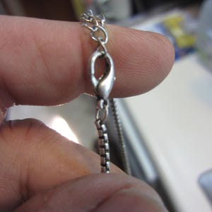 Pu&ograve; includere: Primo piano di una chiusura e catena per collana color argento. La catena ha un design a maglie quadrate. La chiusura &egrave; a forma di artiglio di aragosta. La collana &egrave; tenuta tra le dita.