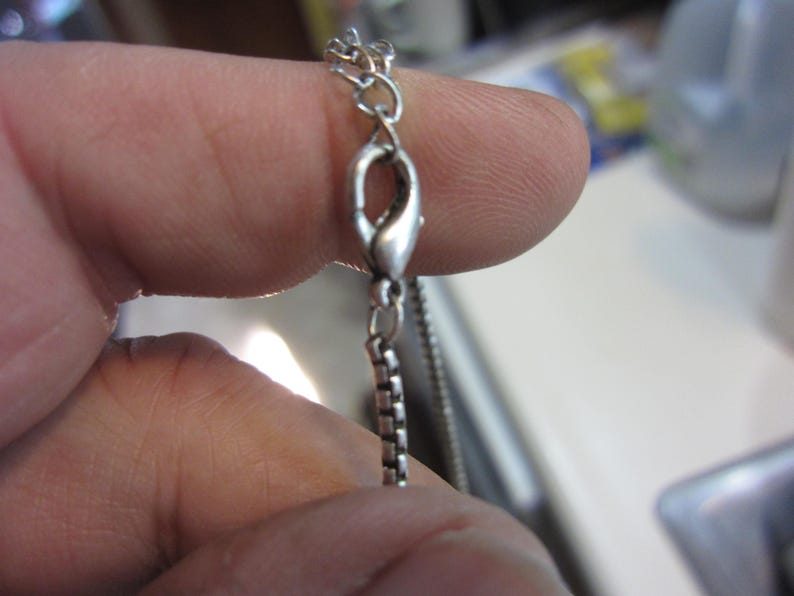 Pu&ograve; includere: Primo piano di una chiusura e catena per collana color argento. La catena ha un design a maglie quadrate. La chiusura &egrave; a forma di artiglio di aragosta. La collana &egrave; tenuta tra le dita.