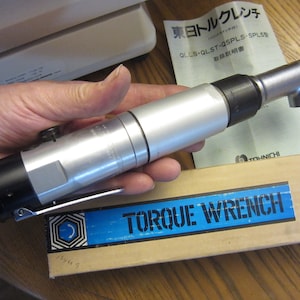 May include: A silver and black torque wrench with a right angle head. The wrench is in a person's hand. The wrench is in its box with a blue label that says "TORQUE WRENCH". The box also has a label with Japanese text.