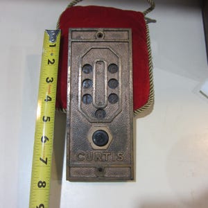 May include: A vintage bronze-colored Curtis door entry system with a series of buttons and the name "CURTIS" at the bottom. The system is approximately 8 inches tall. A red velvet backing is visible.