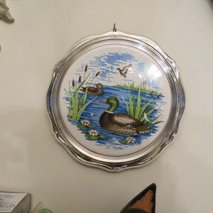 Puede incluir: Un plato redondo de cer&aacute;mica con un marco plateado, que presenta un dise&ntilde;o azul y verde de patos y juncos en un lago.