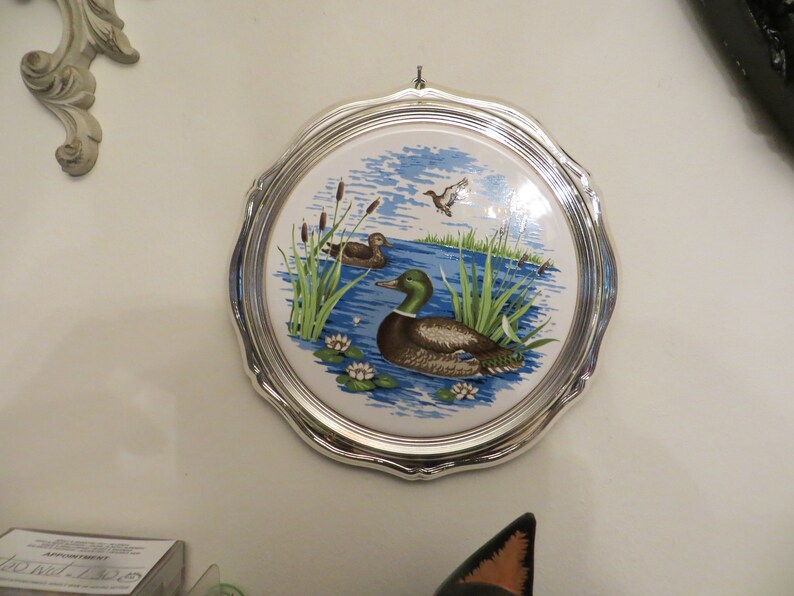 Puede incluir: Un plato redondo de cer&aacute;mica con un marco plateado, que presenta un dise&ntilde;o azul y verde de patos y juncos en un lago.