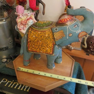 Puede incluir: Una figurita de elefante de madera pintada a mano con detalles azules, amarillos y dorados. El elefante est&aacute; de pie sobre una base de madera con una cinta m&eacute;trica.