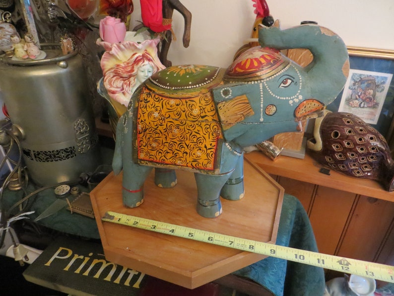 Puede incluir: Una figurita de elefante de madera pintada a mano con detalles azules, amarillos y dorados. El elefante est&aacute; de pie sobre una base de madera con una cinta m&eacute;trica.