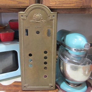 May include: A vintage brass elevator control panel with the words "LIGHT", "ALARM", and "STOP". The panel has several holes and a decorative top. A light blue KitchenAid mixer is to the right.