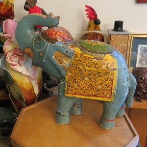 Puede incluir: Una figurita de elefante de madera pintada a mano con colores azul, amarillo y verde. El elefante tiene una silla de montar decorativa con intrincados patrones florales.