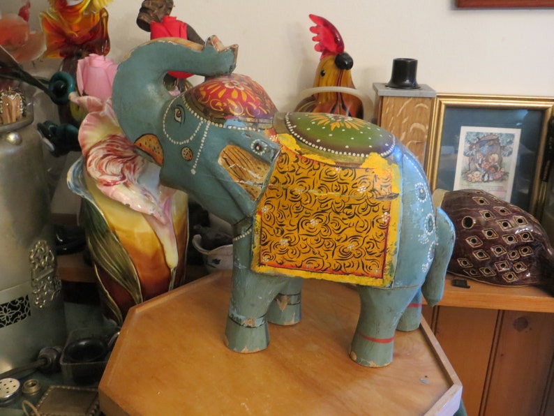 Puede incluir: Una figurita de elefante de madera pintada a mano con colores azul, amarillo y verde. El elefante tiene una silla de montar decorativa con intrincados patrones florales.