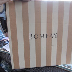 May include: A cardboard box with vertical stripes in alternating shades of beige and white. The word "BOMBAY" is printed in black, centered on the box. The box is held by a person.