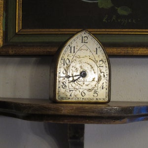 May include: A vintage, silver-toned, arched clock with black hands and Roman numeral markings on a wooden shelf.