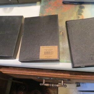 May include: Three black travel journals with different covers. The journal on the left has the word "TRAVEL" embossed on the cover. The journal in the middle has a label that reads "FLEX TRAVEL JOURNAL, STYLE NUMBER 001, BLACK, MADE IN CHINA, 2019". The journal on the right has the words "TRAVEL JOURNAL" embossed on the cover.