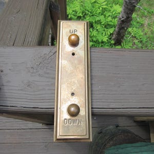 May include: A rectangular brass elevator call button panel with the words "UP" and "DOWN" and two round buttons. The panel is mounted on a wooden surface.