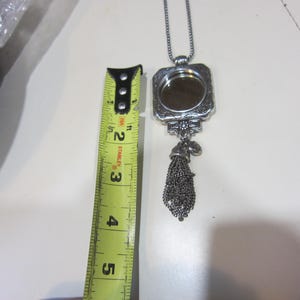 Pu&ograve; includere: Collana con pendente argentato con cornice rettangolare e nappa decorativa. Il pendente &egrave; lungo circa 5 cm. La collana &egrave; su una superficie bianca.