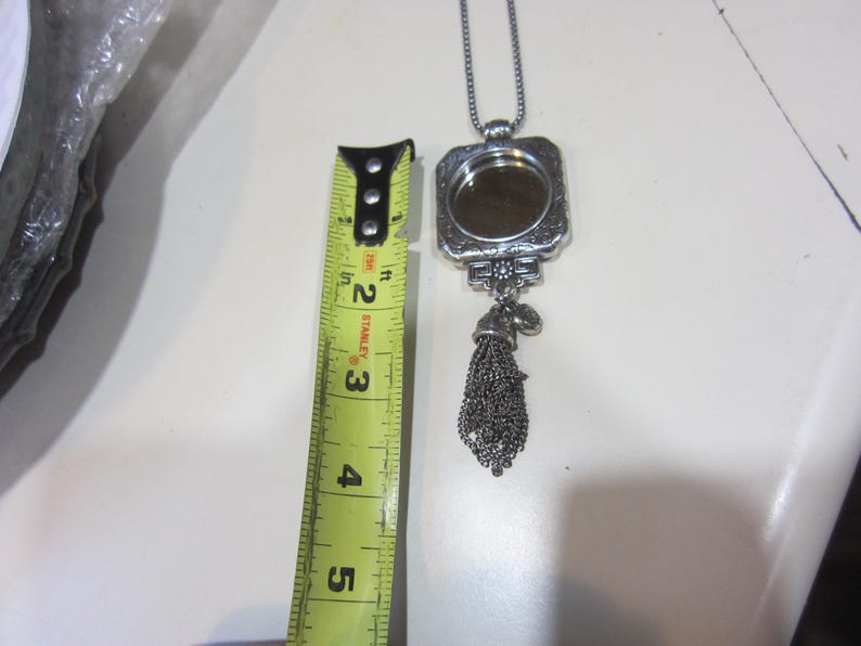 Pu&ograve; includere: Collana con pendente argentato con cornice rettangolare e nappa decorativa. Il pendente &egrave; lungo circa 5 cm. La collana &egrave; su una superficie bianca.