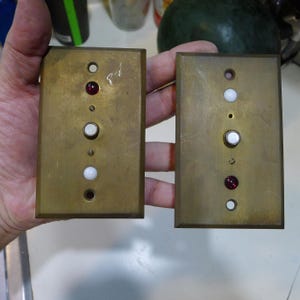 May include: Two rectangular brass wall plates, each featuring a vertical arrangement of three circular components. The left plate has a red jewel, a white button, and a white jewel. The right plate has a white jewel, a white button, and a red jewel.
