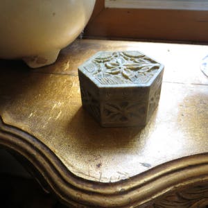 May include: A hexagonal, carved stone box with a floral design. The box is light green and has a detailed pattern on the top and sides. The box is sitting on a wooden surface.