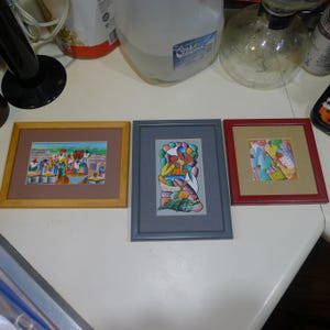 May include: Three framed art prints featuring colorful illustrations of people in outdoor market settings. The left print has a light wood frame, the center print has a gray frame, and the right print has a dark red frame, all with matting.