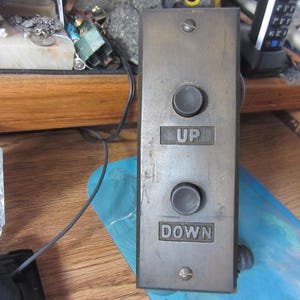 May include: A vintage elevator control panel with two circular buttons and the words "UP" and "DOWN" in block letters. The panel is made of a tarnished metal and has a rectangular shape. The panel is mounted on a wooden surface.