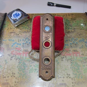 May include: An antique brass elevator call button panel with the words "UP" and "DOWN". The panel has a red velvet backing and a blue light. A silver tape measure and a black flashlight are also visible.