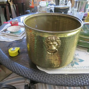 May include: A hammered brass planter with a lion's head ring pull. The planter is cylindrical and has a wide opening. A yellow tape measure is on the table. The planter is approximately 10 inches in diameter and 9 inches tall.
