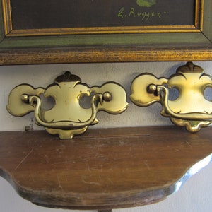 May include: Two ornate brass drawer pulls with a decorative design. The pulls are mounted on a wooden shelf.