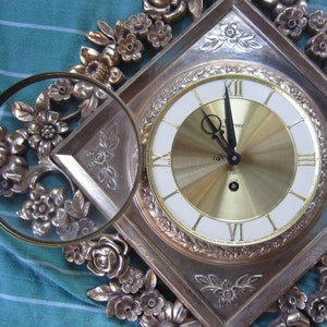 May include: A gold-toned clock with a white face and black hands. The clock is surrounded by a decorative frame with floral designs.