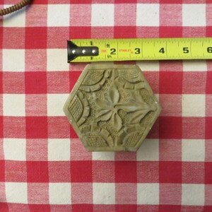 May include: A hexagonal, carved, light brown stone block with a floral design. The block is sitting on a red and white checkered tablecloth.