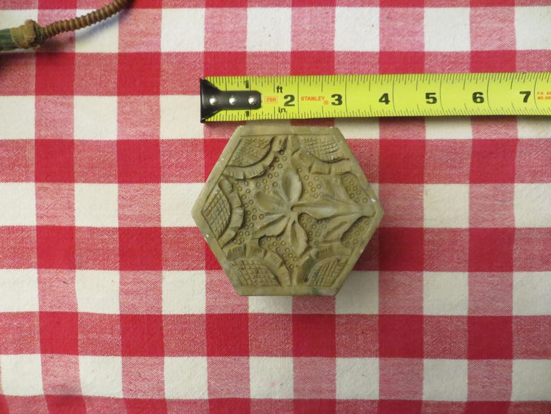 May include: A hexagonal, carved, light brown stone block with a floral design. The block is sitting on a red and white checkered tablecloth.