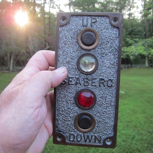 May include: Vintage elevator call button panel with the text "UP" and "DOWN". The panel has a textured surface and four circular buttons, including a red and a clear button. The word "SEABERG" is also visible.