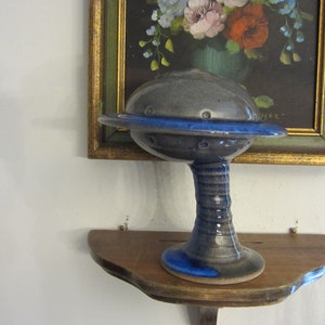 May include: A ceramic serving dish in the shape of a flying saucer with a blue rim and a gray base. The dish is on a wooden shelf.
