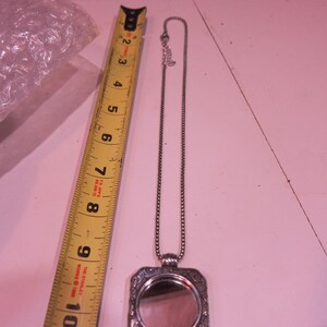 Pu&ograve; includere: Una collana con pendente argentato con una cornice rettangolare ornata e un piccolo specchio. La collana &egrave; accanto a un metro a nastro giallo, che mostra una lunghezza di circa 20 cm.