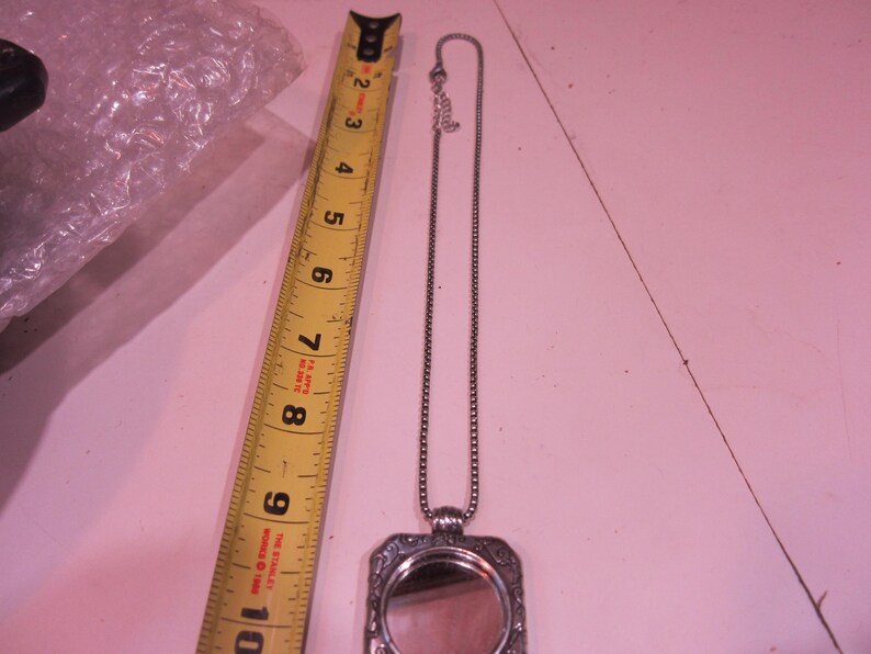 Pu&ograve; includere: Una collana con pendente argentato con una cornice rettangolare ornata e un piccolo specchio. La collana &egrave; accanto a un metro a nastro giallo, che mostra una lunghezza di circa 20 cm.