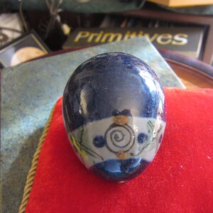 May include: A blue ceramic egg with a white band and a swirl design in brown, yellow, and orange. The egg is sitting on a red velvet surface.