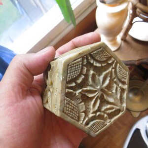 May include: A hand holding a small, intricately carved, hexagonal box made of light green stone. The box has a floral design with raised details.