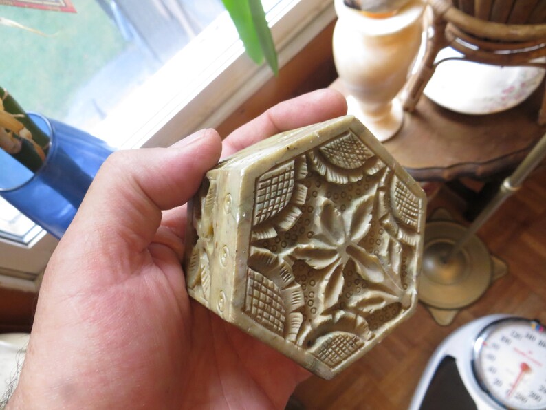 May include: A hand holding a small, intricately carved, hexagonal box made of light green stone. The box has a floral design with raised details.
